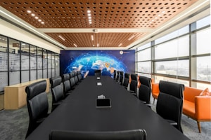 A modern conference room features a long black table surrounded by leather chairs with high backs. On one side, large windows allow natural light to fill the space. The opposite wall displays a mural of a world map with digital network graphics. Orange sofas are placed along the windows, adding a pop of color.