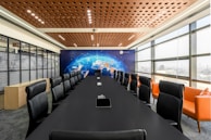 A modern conference room features a long black table surrounded by leather chairs with high backs. On one side, large windows allow natural light to fill the space. The opposite wall displays a mural of a world map with digital network graphics. Orange sofas are placed along the windows, adding a pop of color.