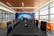 A modern conference room features a long black table surrounded by leather chairs with high backs. On one side, large windows allow natural light to fill the space. The opposite wall displays a mural of a world map with digital network graphics. Orange sofas are placed along the windows, adding a pop of color.