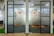 Glass doors with a modern dot pattern design separate an office space. The pattern consists of blue dots of varying sizes, with an orange circular logo positioned near the center of each door. The room beyond features office chairs and orange circular carpets on a dark floor. The interior is well-lit, combining natural and artificial lighting.