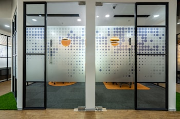 Glass doors with a modern dot pattern design separate an office space. The pattern consists of blue dots of varying sizes, with an orange circular logo positioned near the center of each door. The room beyond features office chairs and orange circular carpets on a dark floor. The interior is well-lit, combining natural and artificial lighting.
