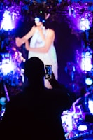 A person is taking a photo with a smartphone in a dark, vibrant setting. The scene includes bright, colorful lights in shades of purple and blue, creating an illuminated atmosphere. The background features a large, blurry image of an individual performing, possibly on stage.