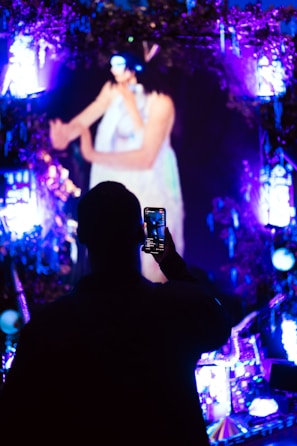 A person is taking a photo with a smartphone in a dark, vibrant setting. The scene includes bright, colorful lights in shades of purple and blue, creating an illuminated atmosphere. The background features a large, blurry image of an individual performing, possibly on stage.