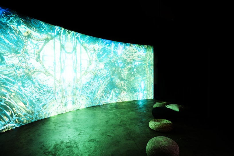 A large, curved screen displays vibrant, abstract patterns with dominant shades of blue and green, creating a surreal and immersive atmosphere. The room is dimly lit, enhancing the luminous effect of the screen. In the foreground, there are several round cushions placed on a concrete floor.