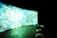 A large, curved screen displays vibrant, abstract patterns with dominant shades of blue and green, creating a surreal and immersive atmosphere. The room is dimly lit, enhancing the luminous effect of the screen. In the foreground, there are several round cushions placed on a concrete floor.