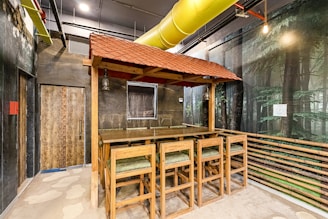 An indoor space featuring a rustic bar setup with wooden tables and chairs under a red-tiled canopy. The area has walls depicting a forest scene, a large yellow ventilation duct running across the ceiling, and exposed pipes. The floor appears to have a stone-like pattern, and there's a closed wooden door to the left.