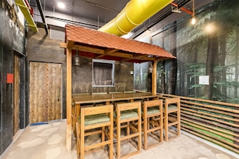 An indoor space featuring a rustic bar setup with wooden tables and chairs under a red-tiled canopy. The area has walls depicting a forest scene, a large yellow ventilation duct running across the ceiling, and exposed pipes. The floor appears to have a stone-like pattern, and there's a closed wooden door to the left.