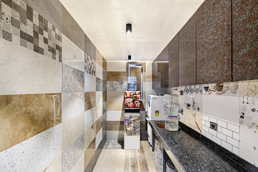 A narrow kitchen space with a patterned wall made of a mix of various tiles displaying intricate geometric and floral designs. The countertops are dark with a glossy finish. On the right side, there is a water dispenser with several water bottles beside it. Cabinets with textured doors line the upper part of the wall. Natural and artificial lighting create a well-lit environment.