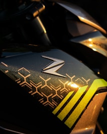 A close-up view of a metallic surface with a distinctive 'Z' logo and geometric patterns in gold and green hues, reflecting light. The surface appears to be part of a vehicle, possibly a motorcycle.