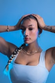 A person with long braided hair is wearing hoop earrings and layered necklaces. They hold their hands on their head, showcasing tattoos on their arm. The background is a gradient of light blue.