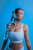 A person with long braided hair and a tattoo on their arm poses against a blue background. They are wearing a white sleeveless top, gold chain necklaces, and earrings, looking to the side.