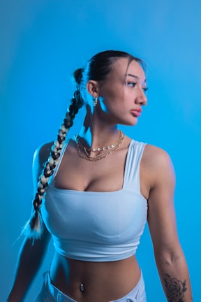 A person with long braided hair and a tattoo on their arm poses against a blue background. They are wearing a white sleeveless top, gold chain necklaces, and earrings, looking to the side.