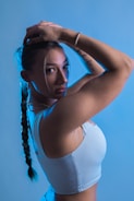 A person with long braided hair is posed with arms raised, creating a dynamic and confident stance. The blue lighting adds a dramatic and modern effect, highlighting the contours of the body and the details of the person's clothing.