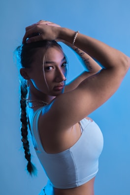 A person with long braided hair is posed with arms raised, creating a dynamic and confident stance. The blue lighting adds a dramatic and modern effect, highlighting the contours of the body and the details of the person's clothing.