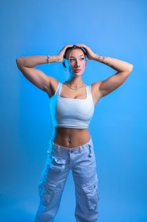 A person posing in front of a plain blue background, wearing a white sleeveless top and white cargo pants. The individual has tattoos on their arms and is accessorized with jewelry including a belly button piercing, bracelets, a necklace, and hoop earrings. Their hands are placed on their head, and they have a focused expression.
