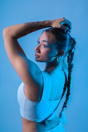 A person with long braided hair wearing a white sleeveless top poses with one hand raised behind their head. They have tattoos on their upper arm and wear small hoop earrings. The background and lighting create a cool blue ambiance.