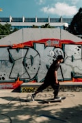 A person is skateboarding in front of a wall covered with colorful graffiti, under a clear blue sky. The graffiti art features large letters and bold colors like red, black, and gray. Shadows from nearby trees are cast on the ground.