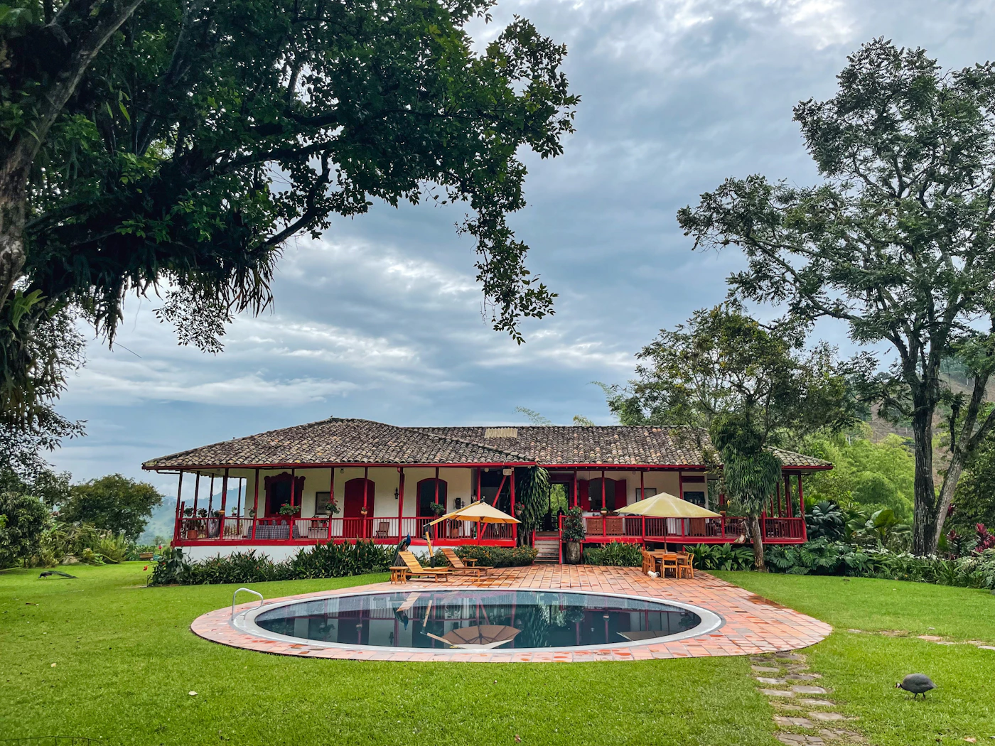 Traditional Colombian hacienda finca estate with red and white colonial architecture and a round circular pool on a green lawn surrounded by tropical trees and Andes mountains behind