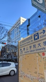 A city scene showing a street map sign indicating directions to Meiji Jingumae Subway Station and Aoyama Gakuin University. A modern glass building is visible in the background with reflections of its surroundings. There are traffic lights and a white car on the road. Bare trees suggest a winter or early spring setting.