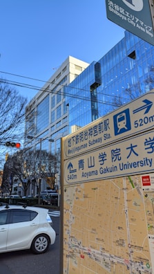 A city scene showing a street map sign indicating directions to Meiji Jingumae Subway Station and Aoyama Gakuin University. A modern glass building is visible in the background with reflections of its surroundings. There are traffic lights and a white car on the road. Bare trees suggest a winter or early spring setting.