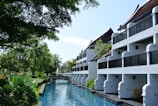 A serene resort setting with white buildings featuring balconies overlooking a tranquil waterway. Lush greenery surrounds the area, with tall palm trees and various tropical plants. The sky is clear, and the atmosphere is peaceful.