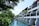 A serene resort setting with white buildings featuring balconies overlooking a tranquil waterway. Lush greenery surrounds the area, with tall palm trees and various tropical plants. The sky is clear, and the atmosphere is peaceful.