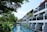 A serene resort setting with white buildings featuring balconies overlooking a tranquil waterway. Lush greenery surrounds the area, with tall palm trees and various tropical plants. The sky is clear, and the atmosphere is peaceful.