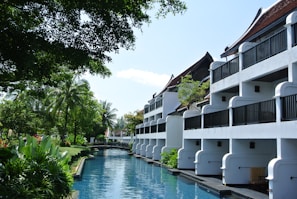 A serene resort setting with white buildings featuring balconies overlooking a tranquil waterway. Lush greenery surrounds the area, with tall palm trees and various tropical plants. The sky is clear, and the atmosphere is peaceful.