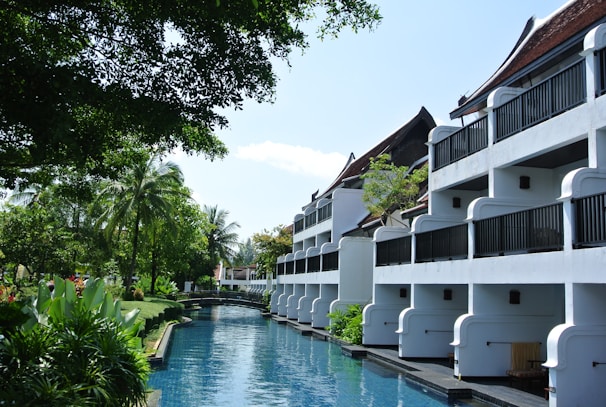 A serene resort setting with white buildings featuring balconies overlooking a tranquil waterway. Lush greenery surrounds the area, with tall palm trees and various tropical plants. The sky is clear, and the atmosphere is peaceful.