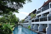 A serene resort setting with white buildings featuring balconies overlooking a tranquil waterway. Lush greenery surrounds the area, with tall palm trees and various tropical plants. The sky is clear, and the atmosphere is peaceful.