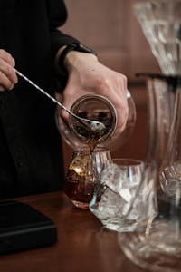 A person is using a stirring rod to mix a beverage being poured from a small glass pitcher into a glass with ice cubes. The scene is set on a wooden surface, accompanied by other glassware and what appears to be a scale.