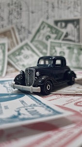 A vintage black toy car is placed on top of various pieces of currency and financial documents in different colors, including green and red. The background is filled with out-of-focus financial paperwork, creating a context of finance or wealth.