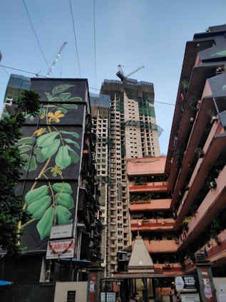 A construction site is visible with a tall building under development, surrounded by residential buildings. The foreground has a building with a large mural depicting green leaves on a dark background. There are cranes on top of the new construction, and some scaffolding and netting are present. The sky is clear with some visible power lines.