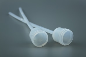Two white plastic lids attached to thin tubes are placed against a soft gray background. The surface texture is smooth, and the absence of shadows gives a minimalistic and sterile appearance.