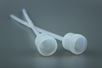 Two white plastic lids attached to thin tubes are placed against a soft gray background. The surface texture is smooth, and the absence of shadows gives a minimalistic and sterile appearance.