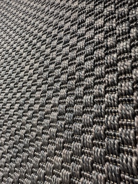 A close-up view of a textured surface with a woven pattern. The image captures a series of interlocking, dark-colored fibers that create a repetitive, geometric design with a slight shine under light.