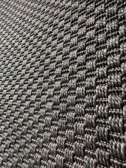 A close-up view of a textured surface with a woven pattern. The image captures a series of interlocking, dark-colored fibers that create a repetitive, geometric design with a slight shine under light.