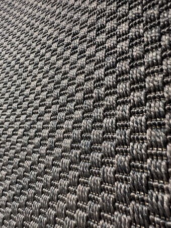 A close-up view of a textured surface with a woven pattern. The image captures a series of interlocking, dark-colored fibers that create a repetitive, geometric design with a slight shine under light.
