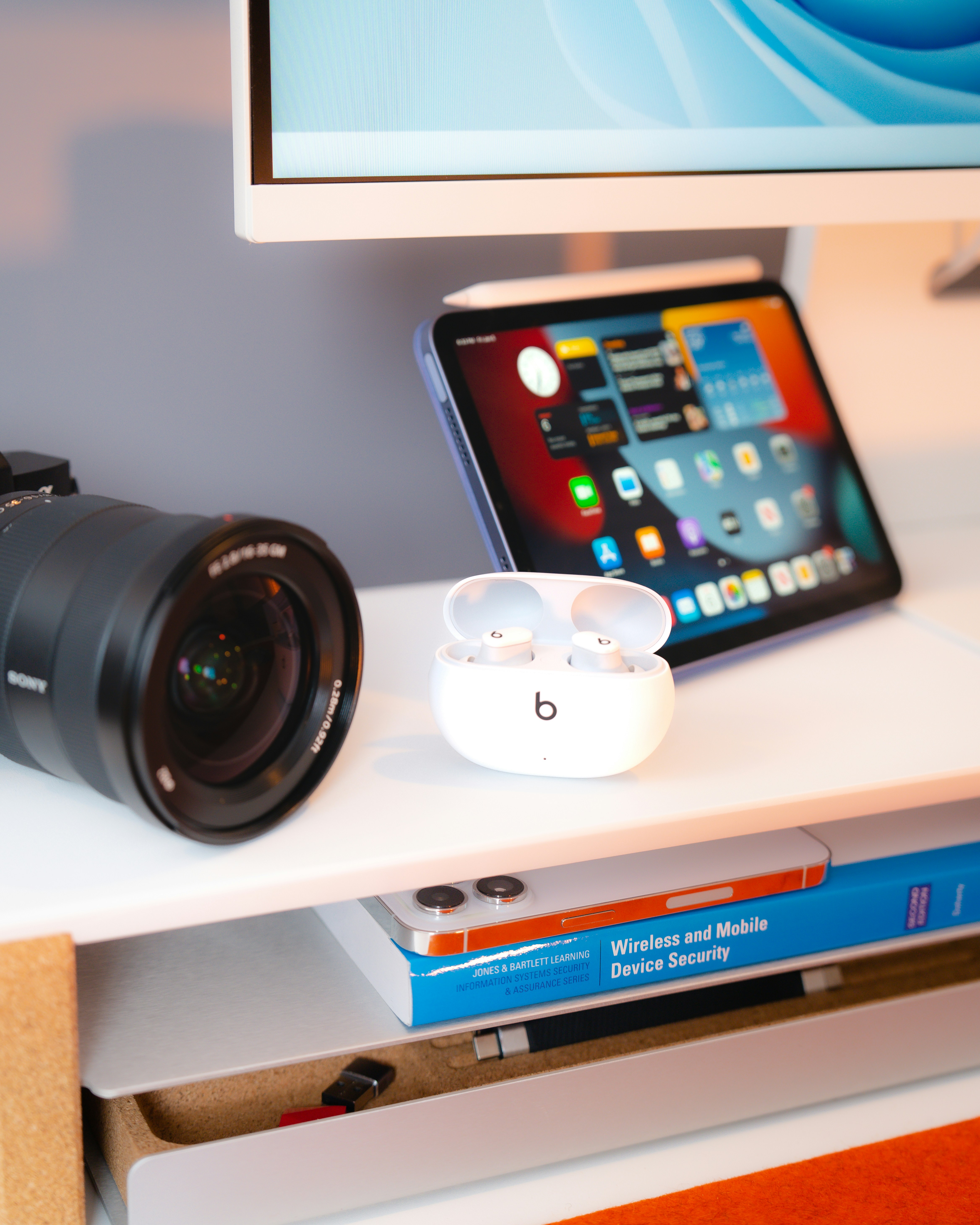 A modern desk setup featuring a camera lens, a tablet displaying an interface with app icons, a pair of earbuds in an open charging case, and a book titled 'Wireless and Mobile Device Security' on a shelf below.