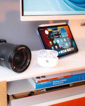 A modern desk setup featuring a camera lens, a tablet displaying an interface with app icons, a pair of earbuds in an open charging case, and a book titled 'Wireless and Mobile Device Security' on a shelf below.
