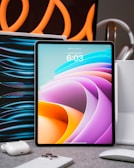A tablet with a colorful abstract display is placed upright on a table. Surrounding it are various tech accessories, including a smartphone, earbuds, and a mouse. Curved orange lines in the background create an artistic flair, suggesting a modern and high-tech setting.