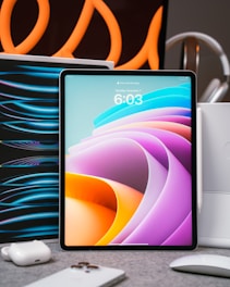 A tablet with a colorful abstract display is placed upright on a table. Surrounding it are various tech accessories, including a smartphone, earbuds, and a mouse. Curved orange lines in the background create an artistic flair, suggesting a modern and high-tech setting.