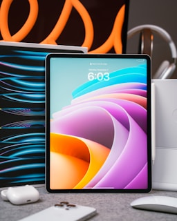 A tablet with a colorful abstract display is placed upright on a table. Surrounding it are various tech accessories, including a smartphone, earbuds, and a mouse. Curved orange lines in the background create an artistic flair, suggesting a modern and high-tech setting.