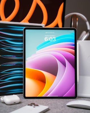 A tablet with a colorful abstract display is placed upright on a table. Surrounding it are various tech accessories, including a smartphone, earbuds, and a mouse. Curved orange lines in the background create an artistic flair, suggesting a modern and high-tech setting.