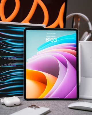 A tablet with a colorful abstract display is placed upright on a table. Surrounding it are various tech accessories, including a smartphone, earbuds, and a mouse. Curved orange lines in the background create an artistic flair, suggesting a modern and high-tech setting.