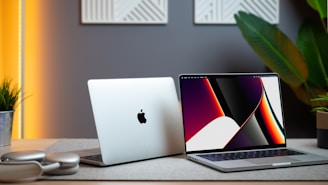 Two laptops are positioned on a desk with modern decor. One laptop is open, displaying a colorful abstract design on the screen, while the other is closed, showing an apple logo. The setup is accompanied by a pair of headphones, a potted plant, and warm ambient lighting, creating a sophisticated workspace.