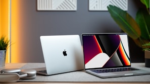 Two laptops are positioned on a desk with modern decor. One laptop is open, displaying a colorful abstract design on the screen, while the other is closed, showing an apple logo. The setup is accompanied by a pair of headphones, a potted plant, and warm ambient lighting, creating a sophisticated workspace.