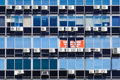 Rows of rectangular windows on a building facade, each equipped with air conditioning units. A sign with text in Spanish appears on one of the windows, indicating a space for rent. The windows vary in shades of blue, adding a uniform and repetitive architectural pattern.