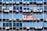 Rows of rectangular windows on a building facade, each equipped with air conditioning units. A sign with text in Spanish appears on one of the windows, indicating a space for rent. The windows vary in shades of blue, adding a uniform and repetitive architectural pattern.