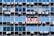 Rows of rectangular windows on a building facade, each equipped with air conditioning units. A sign with text in Spanish appears on one of the windows, indicating a space for rent. The windows vary in shades of blue, adding a uniform and repetitive architectural pattern.
